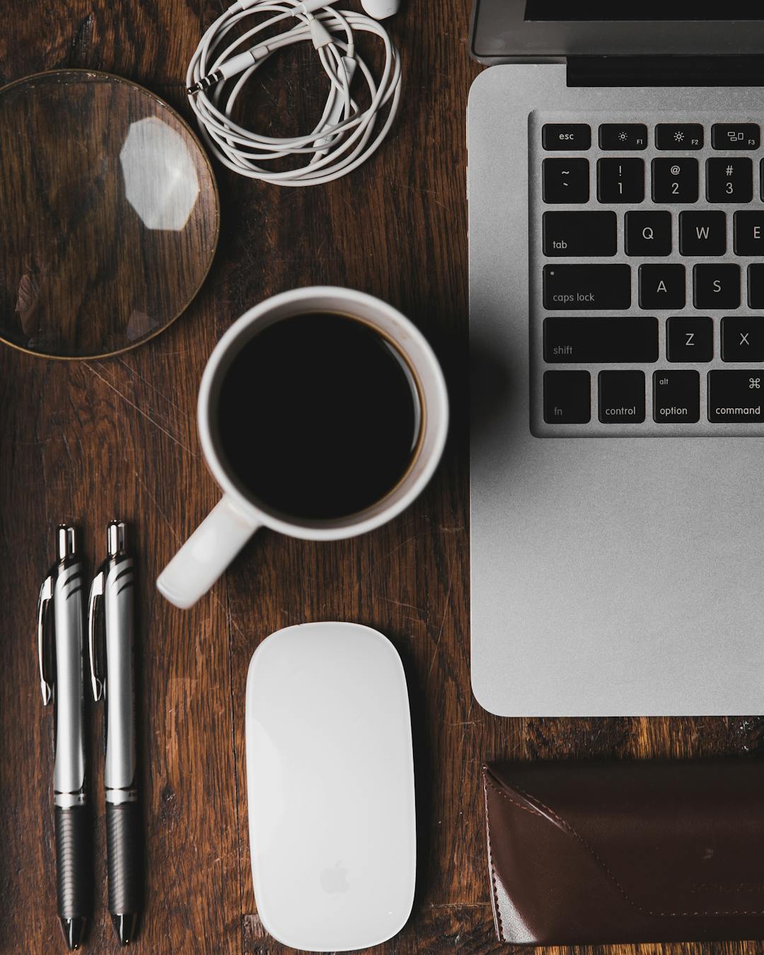 A laptop, a cup of coffee, a mouse and two pens on a wooden desk, seen from above